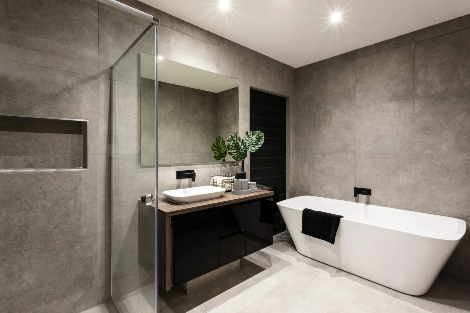 Modern bathroom with a shower area and bath tub including a wall mirror beside a fancy plant near a tap and sink over the wooden counter and dark cupboard